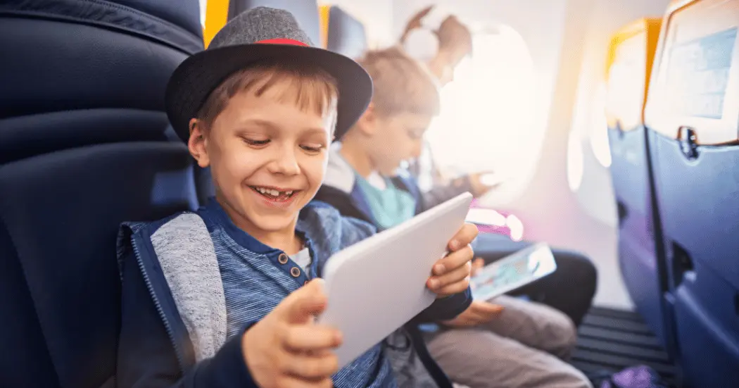 A young boy enjoys entertainment on a tablet while flying in an airplane, his brothers sits next to him holding his own tablet.