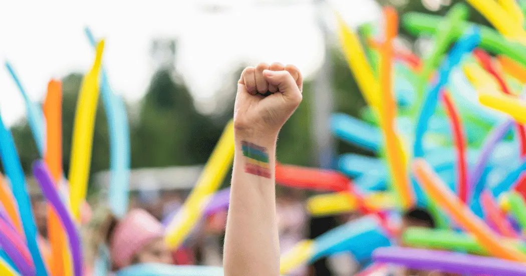 At Pride celebration, we see a raised hand in focus. The person's wrist shows a rainbow-colored drawing.
