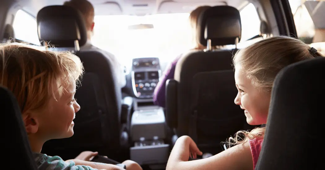 Two kids sitting in the back seats of a car smile at each other while their parents navigate a road trip.