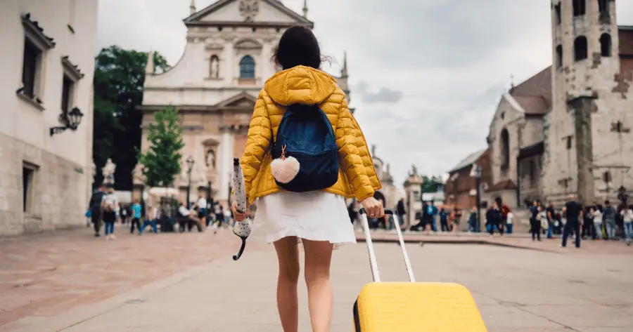 A woman arrives at her solo travel destination, carrying a suitcase, umbrella, and backpack